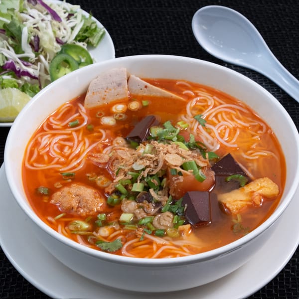 A vibrant bowl of Vietnamese bún bò Huế soup with noodles, meat, and fresh herbs, served with a side salad.