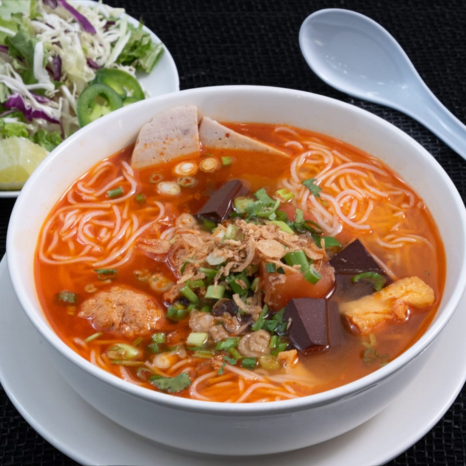 A vibrant bowl of Vietnamese bún bò Huế soup with noodles, meat, and fresh herbs, served with a side salad.