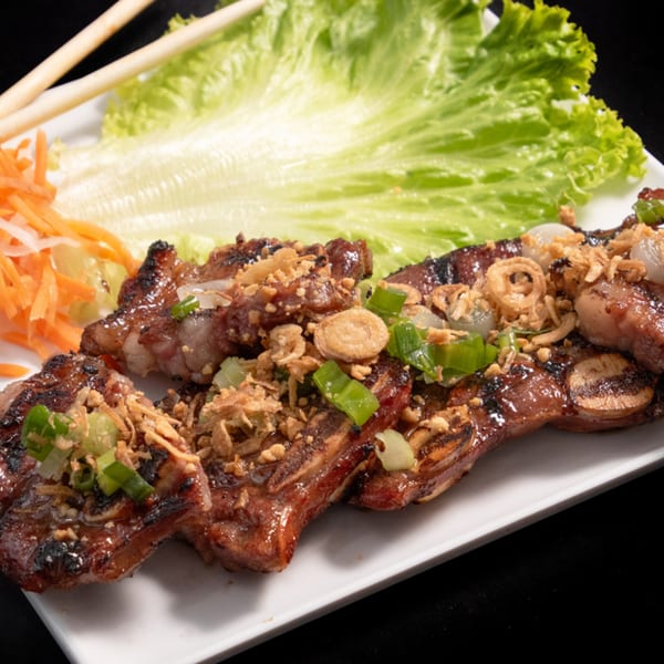 Grilled Vietnamese short ribs with fried shallots, green onions, lettuce, and pickled carrots on a white plate.