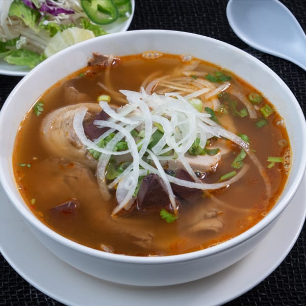 A bowl of Vietnamese Phở Skyline soup with meat, noodles, onions, and herbs, with a side of vegetables.