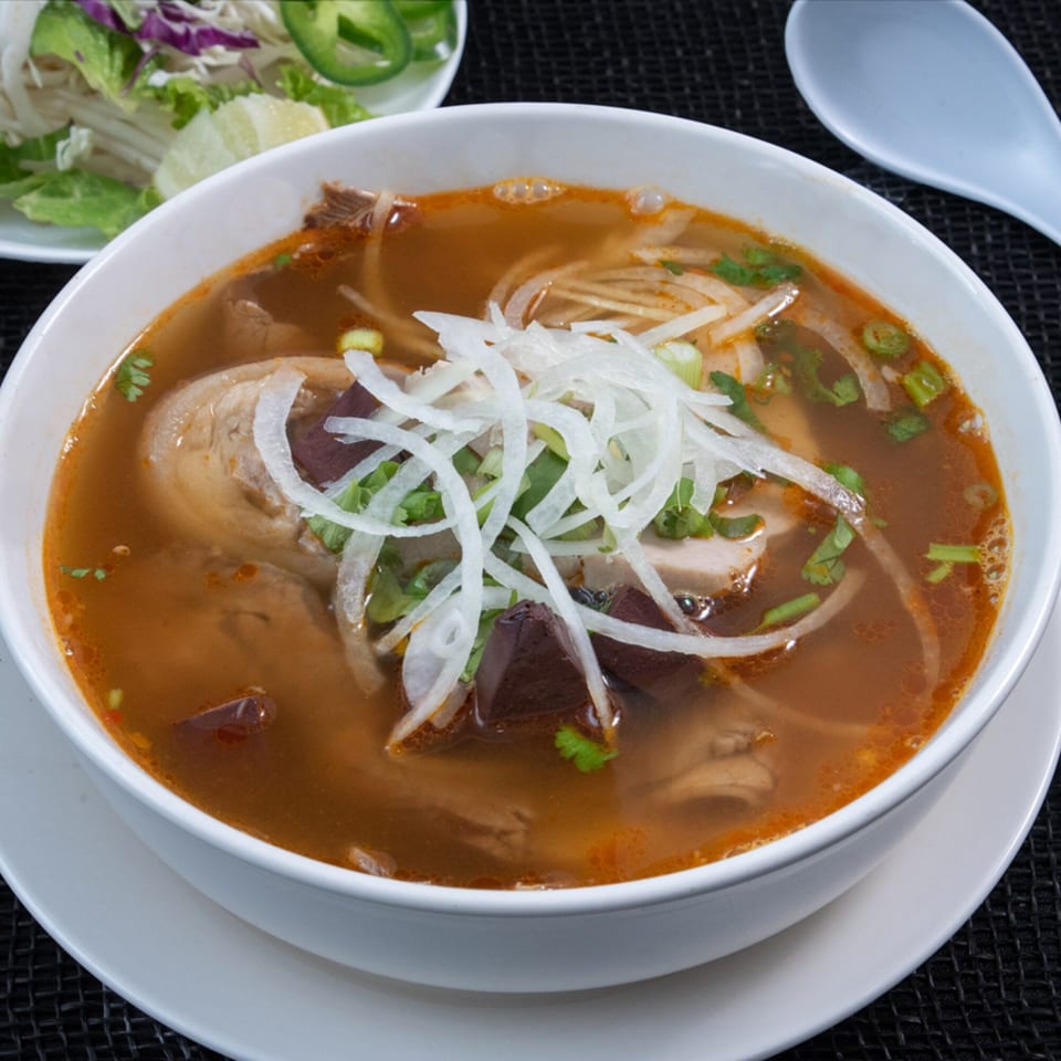 A bowl of Vietnamese Phở Skyline soup with meat, noodles, onions, and herbs, with a side of vegetables.