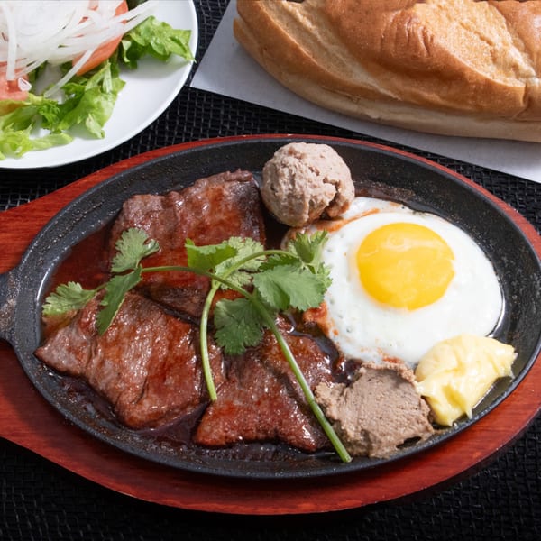 Sizzling Vietnamese steak and eggs with pate, cilantro, and a side of baguette and salad.